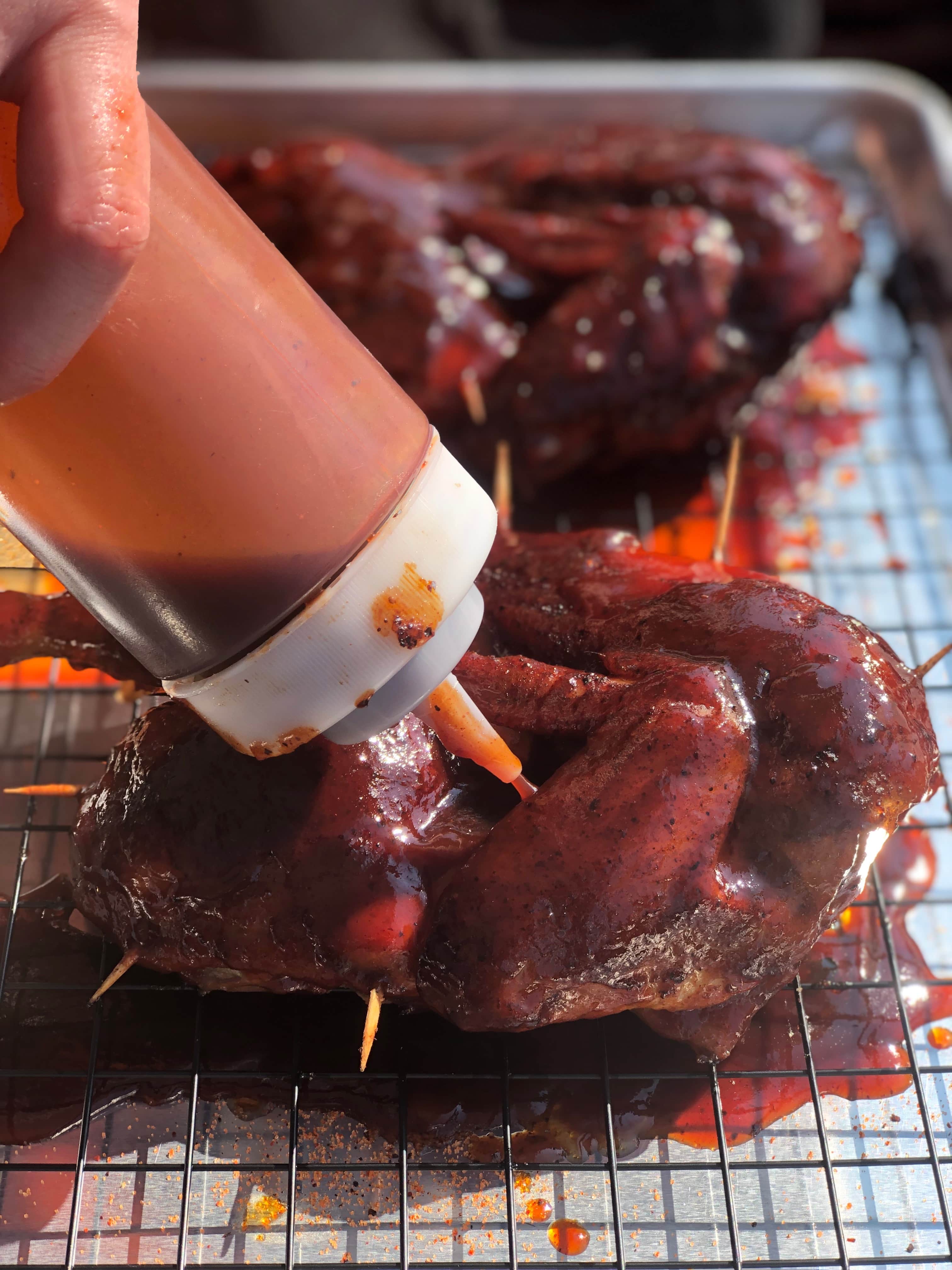 Barbecue sauce being applied to seasoned smoked chicken on a wire rack.