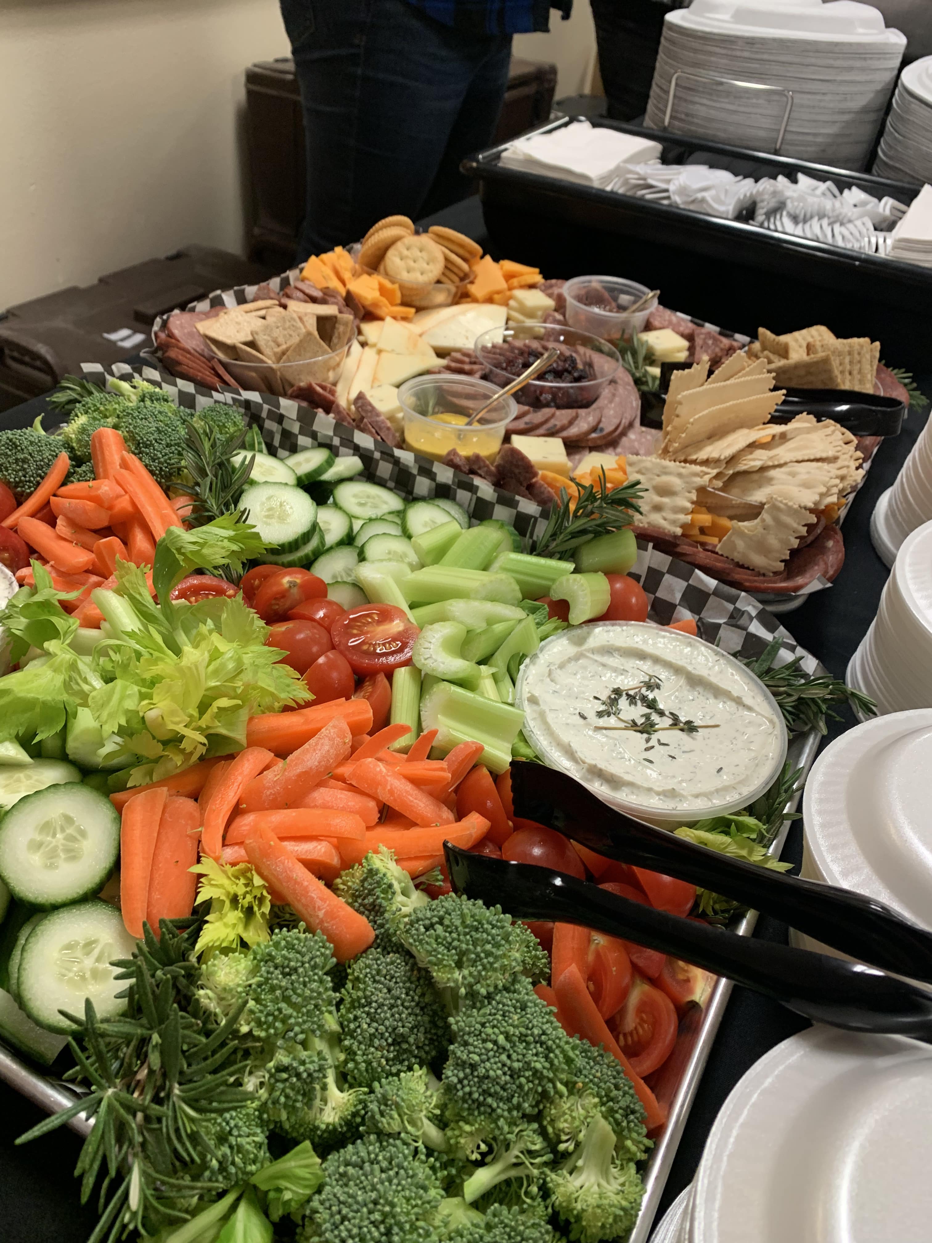 Catering spread with fresh vegetables, herb dip, sliced meats, cheeses, crackers, and condiments.