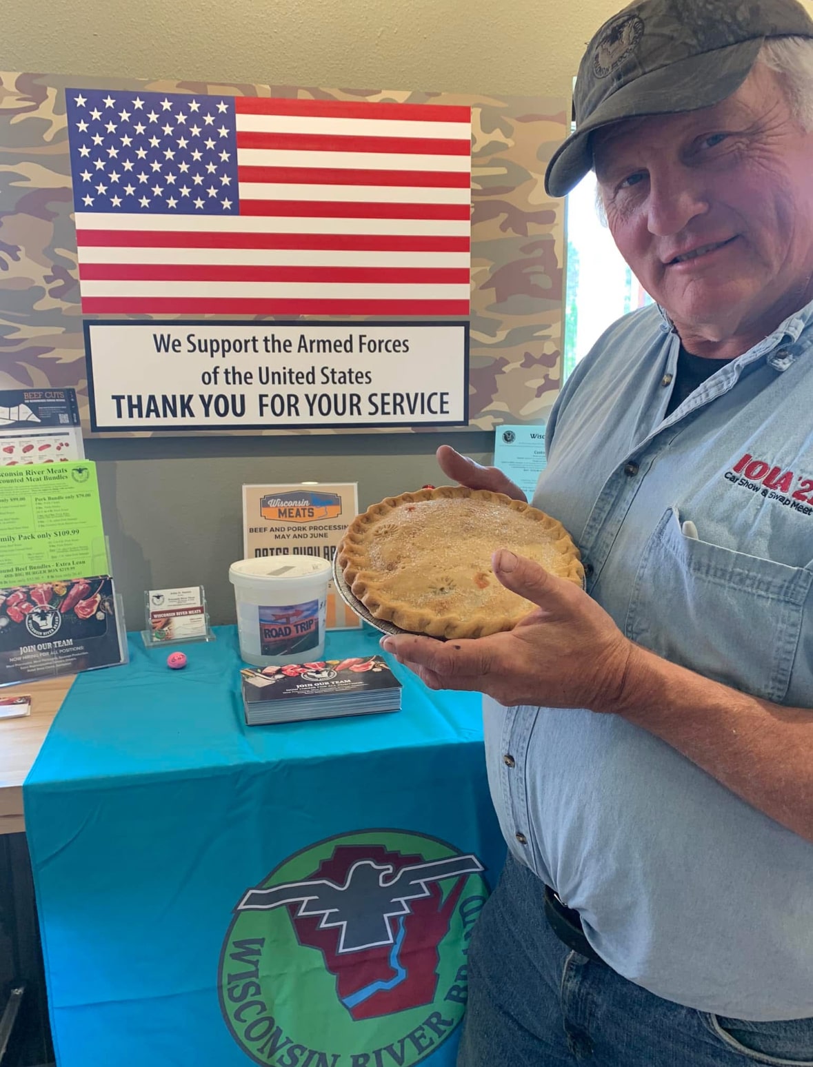 John Hamm, President of Wisconsin River Meats, holding a pie beside an American flag and a sign thanking members of the U.S. Armed Forces.