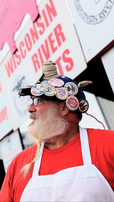 Wisconsin River Meats butcher with a white beard wearing a red shirt, white apron, and hat decorated with award pins.