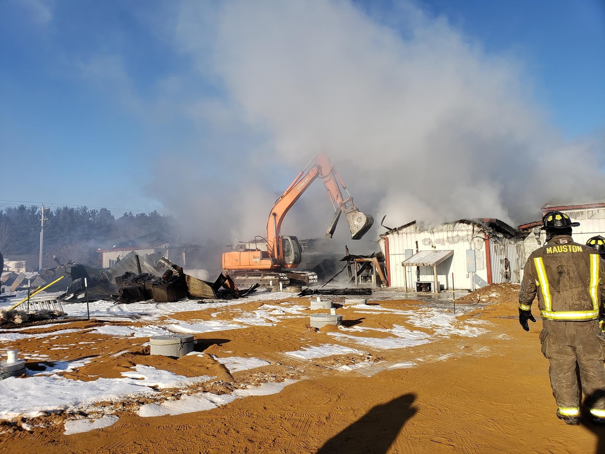 Excavator removing fire-damaged sections of the Wisconsin River Meats facility as firefighters monitor heavy smoke in winter.